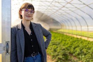 A photo of a white woman with brown hair and glasses standing in a greenhouse. There are plants growing in the background. She is wearing a gray blazer, black blouse, and jeans.